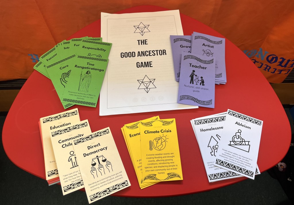 Printed copies of The Good Ancestor Game guidebook and card decks are displayed on a red table